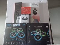 Photo showing two smart watches, two earbuds boxes, and two drone boxes all stacked together on a white surface with visible labels and packaging.