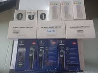 Photo showing all items arranged in rows including fitness trackers in small white boxes, Aqara night lights labeled boxes, in-wall smart switches in white larger rectangular boxes, and Ceenwes branded hair clippers in blue boxes.