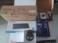 Photo showing boxed RENPHO cordless massager, OMOTON wireless keyboard, battery charger in foam packaging, Charmsman beard trimmer box, and hearing aid device in clear container, all placed on a grey surface.