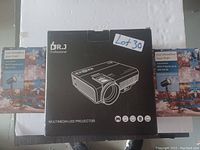 Photo showing one black box of Dr.J Multimedia LED Projector flanked by two colorful boxes of Cool White & Blue Snowflake Projectors on a white background.