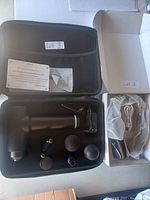 Photo of open black case containing a massage gun with five interchangeable heads and charging cable, plus a gray pouch containing a black wireless neck fan with charging cable.