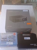 Photo showing the Apeman LC350 projector box, Mini Game Anniversary Edition boxed console, and black carrying case for TENS 3000 machine, all labeled Lot 1.
