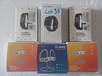 Photo of 3 FKANT Fitness Trackers, 1 Smart Watch, and 3 pairs of wireless sports earphones in their retail packaging, displayed against a white wall.