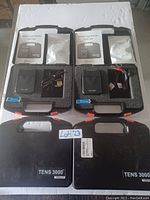 Photo showing two open TENS 3000 cases with electronic stimulation units, manuals, electrode pads, batteries, and cables inside, and two closed cases labeled 'TENS 3000' on table.