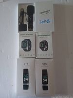 Photo showing all 5 wearables: 2 V19 Smart Bracelets in retail boxes, 2 Smart Bracelets in retail boxes, and 1 black Smart Watch with charging cable outside of boxes.