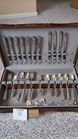Full view of opened wooden box with organized silverware pieces inside holder slots, showing variety of forks and spoons with metal handles.