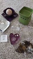 Photo of candle holders and candle melt including green ceramic melt warmer, purple candle holder with beige candle, clear and purple glass decorative holders, and silver heart shaped holder on carpet.