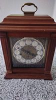 Front view of Hermle mechanical clock with cherry wood case, brass handle, and ornate clock face with three winding holes.