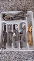 Tray with stainless steel forks, spoons, knives with ornate handles in organized sections, also some gold colored flatware pieces separated