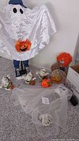 Photo showing white satin-like ghost youth figure with pumpkin decoration, red-haired clown doll, Halloween pumpkin ceramic pieces, and sheer ghost veil with skeletal hand
