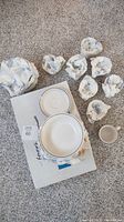 All 24 pieces of the dinnerware set shown in crumpled paper packaging on carpet. Includes bowls, cups, and saucers with visible blue rim pattern.