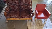 Red vinyl covered small rocking chair with visible signs of wear, placed on a cloth, next to a wooden bench rocker with patterned cushions.