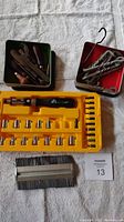 Full view of the lot showing leather working tools in green tin, metal hooks in red tin, socket set with screwdriver and sockets in yellow molded tray, and a metal brush tool.