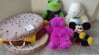 Photo showing all five stuffed items including hamburger plush, green frog, white rabbit, bright pink teddy bear, and Disney Mickey Mouse plush toy.