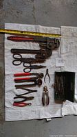 Tools arranged on white cloth including pipe wrenches, tin snips, cutters, pliers, small metal box of bits, and measuring tape showing length.