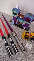 Three light sabers lying next to the Matchbox Haunted House playset and a small yellow toy dozer on carpeted floor.