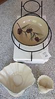 Ceramic bowl with pinecone design on metal stand and three cream seashell-shaped ceramic pieces on carpet.