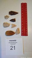 Top down photo of seven arrowheads next to a red ruler for scale and MaxSold lot number 21