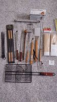 Display of various grilling tools including spatulas, forks, meat presses, grilling basket, metal skewers and packs of wooden skewers.