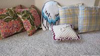 Six pillows displayed on floor including floral, geometric, knitted, and graphic designs