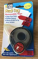 Close-up of a single Redi-Tag dispenser with red 'SIGN HERE' removable tags on a roll inside original packaging.