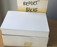 Stacked gray report back papers inside an open box with label 'REPORT BACKS' visible in the background.