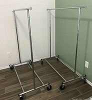 Two silver collapsible garment racks with adjustable height, standing on wooden floor against white and green walls.