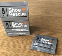 Photo showing a box of Shoe Rescue towelettes and one individual towelette sachet, gray packaging with branding and product use description.