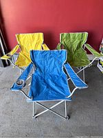 Three camping chairs: yellow, green, and blue, positioned in front of a red wall, showing full seating and armrests with cup holders