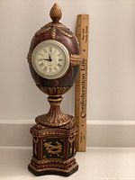 Front view of the full ornate egg-shaped clock with gold leaf decorations on an octagonal base, showing size comparison with a ruler.