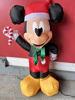 Photo of the fully inflated Mickey Mouse Christmas decoration in front of a red wall. Mickey wears a red Santa hat, green scarf, red shorts, yellow shoes, and holds a candy cane.