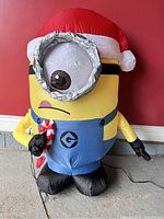 Photo of the inflated Carl Minion character in Santa hat, scarf, overalls, standing against red wall.