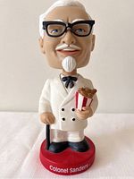Front view of Colonel Sanders KFC Funko bobblehead showing white suit, black glasses, and holding bucket of chicken.