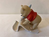 Side view of Winnie The Pooh ceramic figure wearing a red shirt, writing on a white scroll with a blue quill pen.