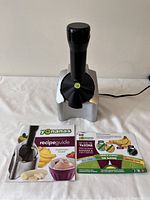 Front view of Yonanas frozen fruit soft serve machine with plunger attached, next to the colorful recipe guide booklet.