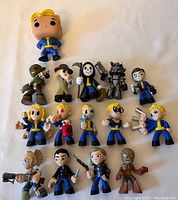 Photo showing 15 Fallout-themed Funko figures including 14 mini figures and 1 Funko Pop Vault Boy figure in blue and yellow outfit, with various poses and accessories.