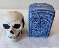 Photo showing two Halloween-themed ceramic salt and pepper shakers, one shaped like a skull and one like an RIP tombstone, on a white surface.