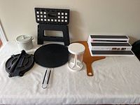 Seven kitchen items arranged on white cloth including tortilla press, ceramic dip server, burger press, splatter guard, foldable step stool, pizza peel, and non-skid shelf organizer