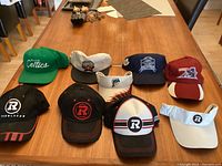 Top-down photo of nine sports hats arranged on a rectangular wooden table, showing various team logos and colors.