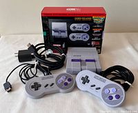 Front view of SNES Classic Mini console with two wired controllers and cables. Original box behind.