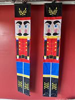Pair of outdoor nutcracker-themed hanging banners photographed against a red wall, showing colors and design details.