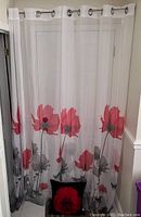 Photo showing two sheer curtains with poppy flower design hung on a door with metal grommets, along with a black pillow featuring a large red poppy embroidered on it placed at the base.