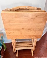 Front view of four wooden TV snack tables stacked together on the stand, showing rectangular tabletops and foldable legs.