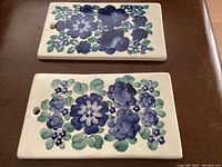 Top view of two rectangular vintage wall plaques with blue floral designs laid flat on a wood surface.