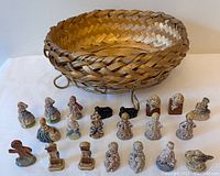 Wide shot of all 21 ceramic Wade England figurines arranged in front of and inside the woven basket. The figures show a variety of characters mainly girls in dresses, some animals and decorative elements.
