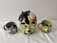 Zombie ceramic cookie jar with detachable lid and two matching coffee cups all featuring detailed zombie faces and colorations.