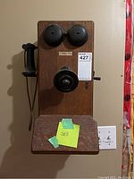 Front view of entire wooden wall‐mount telephone including dual bells, mouthpiece, handset, writing shelf and lot tag
