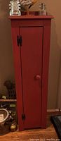 Front view of tall narrow red wooden cabinet with a single door and black metal hardware.