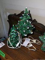 Two green ceramic Christmas trees placed on a wooden table, one smaller (9 inch) and one larger (13 inch) with multiple colored bulbs and white cords with switches