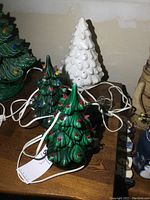 Three ceramic Christmas trees on a wooden surface: two green and lit with multicolored bulbs and one white unfinished tree with an extra base beside them. Visible power cords for plugging in.